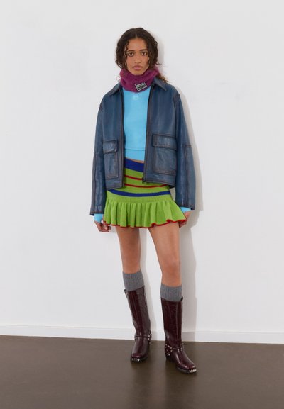 Chaqueta de cuero azul sobre un suéter azul claro, falda de punto multicolor con volantes, calcetas grises hasta la rodilla y botas burdeos hasta la rodilla.