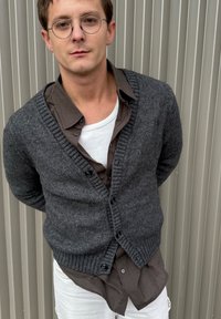 Mørkegrå strikket cardigan med V-hals, ribbede manchetter og knapper, båret over en brunternet skjorte og en hvid undertrøje.