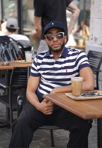 Camisa polo a rayas en azul marino y blanco, pantalones negros. Gorra negra, gafas de sol grandes. Vaso de café helado sobre una mesa de madera. Entorno al aire libre.