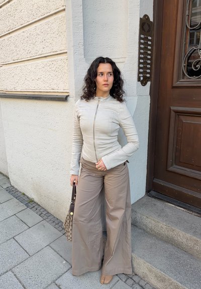 Mujer con cabello rizado oscuro, con chaqueta beige y pantalones anchos color coñac, de pie cerca de la entrada de un edificio, sosteniendo un bolso con estampado, mirando hacia un lado.