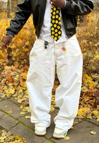 Calças brancas largas, casaco bomber de couro preto, camisa branca às riscas, gravata amarela com smiley, e acessórios de porta-chaves multicoloridos; ténis castanhos.