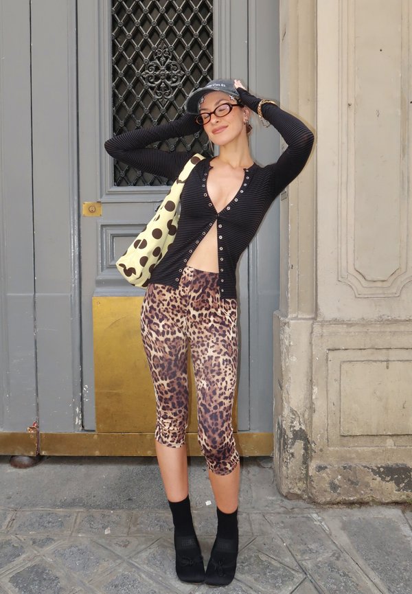 Ung kvinde i leopardmønstrede capri leggings og sort knap-top poserer med hænderne på hovedet og bærer en prikket taske ved en dekorativ dør.