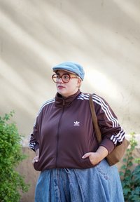 Brun zip-up jakke med hvide striber, lyseblå kasket, oversized denimbukser og store runde briller, parret med en brun skuldertaske.