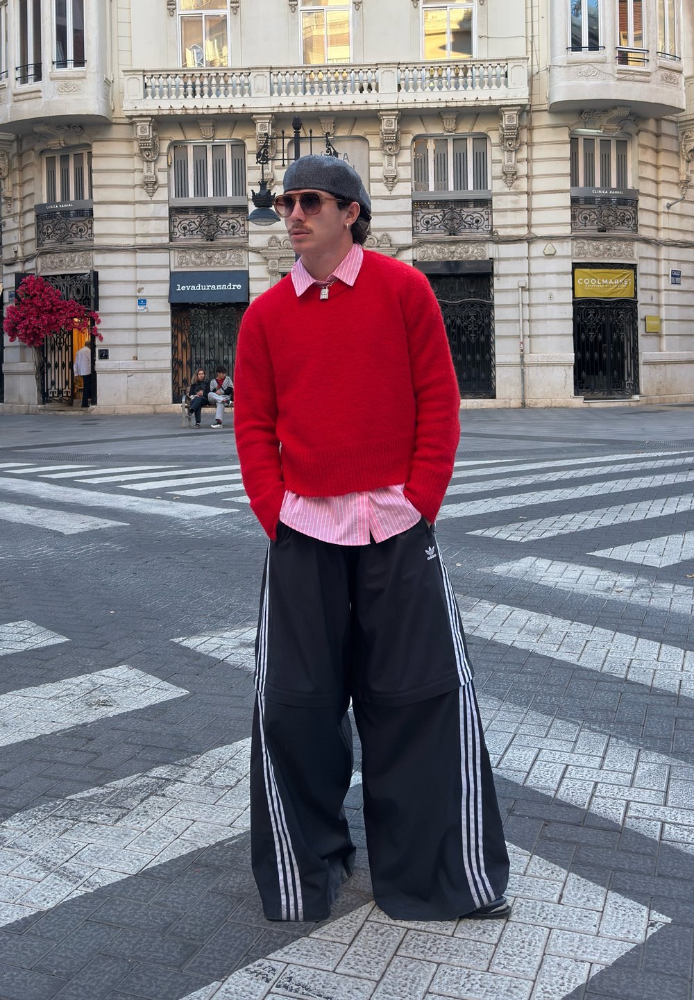 Maglione rosso a maglia sopra a una camicia rosa a righe, abbinato a pantaloni sportivi neri oversize con strisce bianche. Ambientazione urbana di strada.