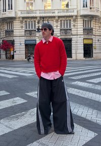 Maglione rosso a maglia sopra a una camicia rosa a righe, abbinato a pantaloni sportivi neri oversize con strisce bianche. Ambientazione urbana di strada.