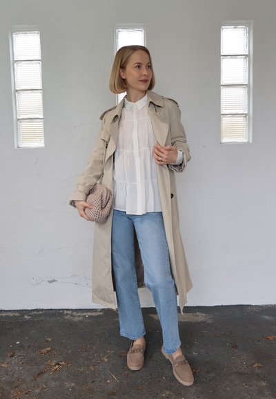 Mujer de pie en el interior vistiendo un abrigo beige, blusa blanca, vaqueros azul claro, mocasines beige y sosteniendo un bolso de mano de punto.