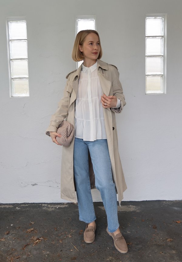 Frau steht drinnen, trägt einen beigen Trenchcoat, eine weiße Bluse, hellblaue Jeans, beige Loafers und hält eine gestrickte Clutch.