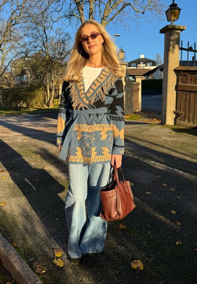 Cárdigan con estampado azul y negro con flecos, pantalones de mezclilla acampanados de talle alto y un bolso de cuero marrón. Gafas de sol completan el conjunto.