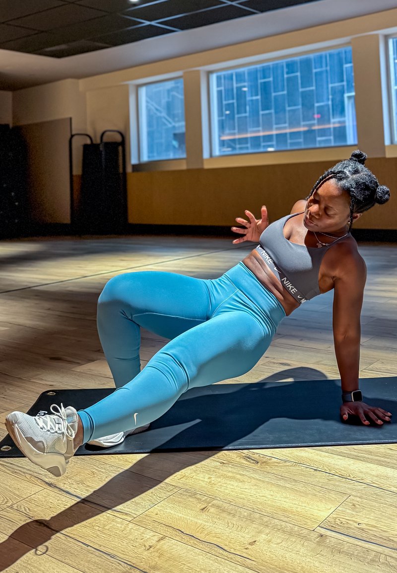 Una persona esegue un esercizio su un tappetino nero, indossando un reggiseno sportivo grigio e leggings blu, con scarpe leggere e finestre sullo sfondo.