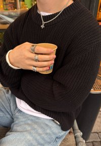 Črn pleten pulover z rebrasto teksturo, prekrit nad belim srajcem. Srebrni prstani in necklace z zvezdnim obešalnikom. Drži kartonsko skodelico.