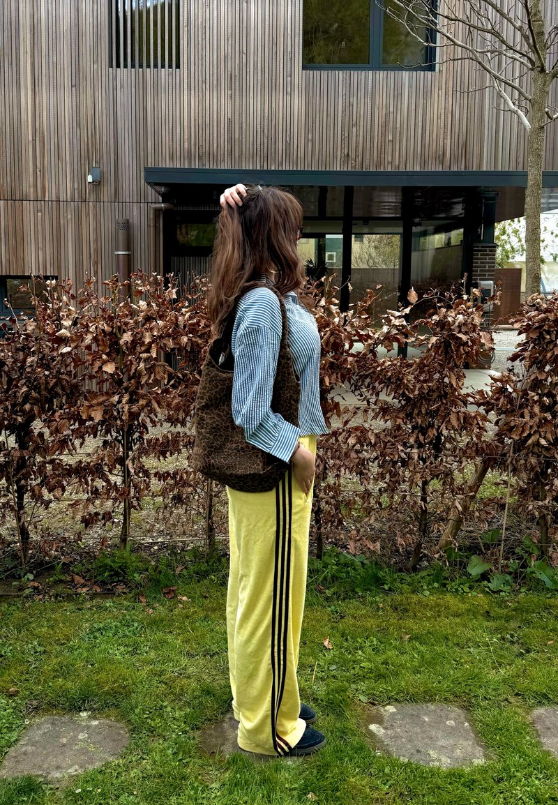 Jonge vrouw staand zijwaarts op het gras, gekleed in gele broek met zwarte strepen, blauw gestreept shirt met lange mouwen en draagt een shopper met luipaardprint.