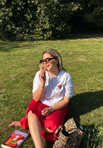 Camiseta blanca con un diseño de corazón rojo, falda de satén roja y gafas de sol grandes negras. Sentada en hierba verde con un bolso de estampado de leopardo y un libro.