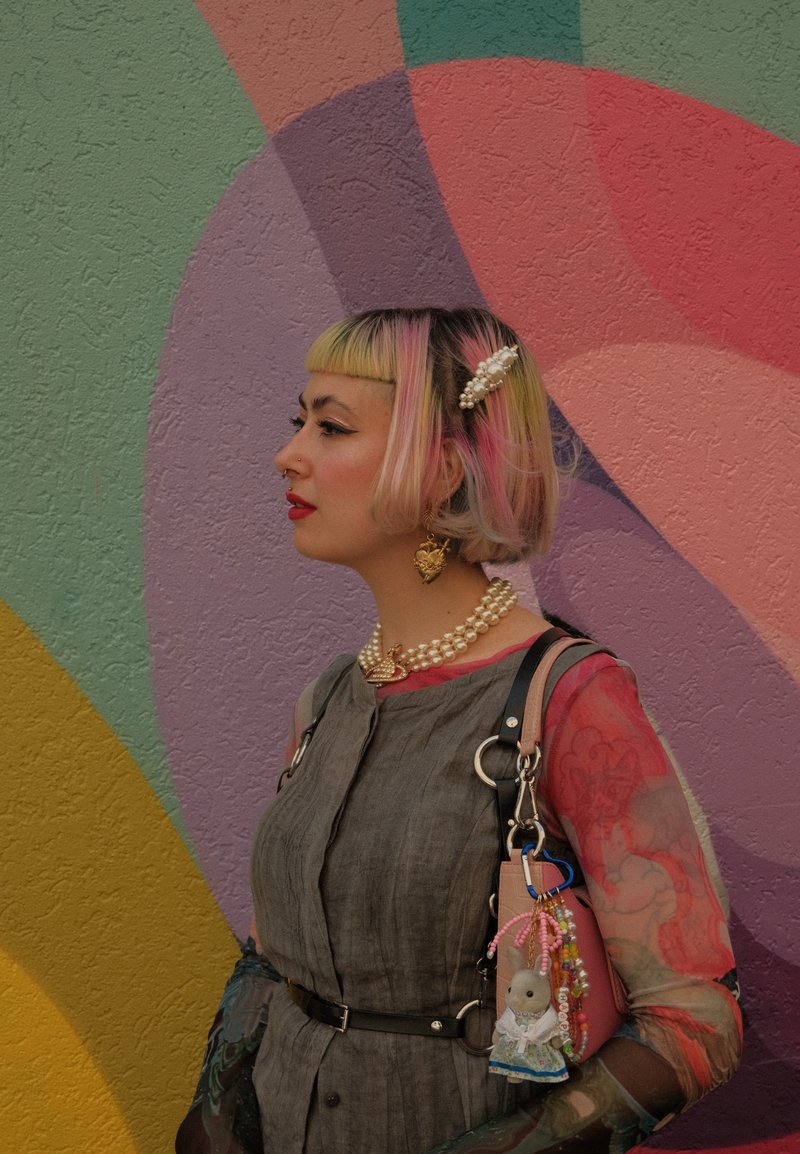 The model is wearing a grey dress cinched with a belt, complemented by pearl jewellery and a bag embellished with charms. The background showcases vibrant abstract patterns.