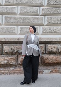 Femme portant un hijab noir, un blazer gris avec une ceinture en dentelle blanche, un col roulé blanc, un pantalon large noir à fines rayures, debout contre un mur en pierre texturé.