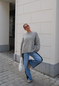 Lys grå, fluffy genser, blå vide jeans med grønne sidestriper, og beige sko. Tilbehøret inkluderer et flerfarget halskjede og briller.