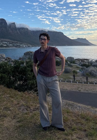 Hombre con gafas, vestido con una camisa morada y pantalones grises, de pie sobre el césped cerca de una costa rocosa con montañas y un cielo parcialmente nublado.