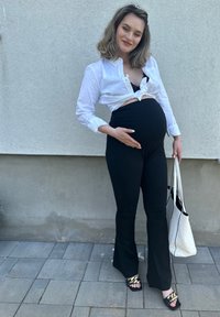 Μαύρο φαρδύ παντελόνι εγκυμοσύνης, λευκό πουκάμισο με κόμπο και μαύρες σανδάλες με λουριά και χρυσές λεπτομέρειες. Κρατώντας μια άσπρη τσάντα σε γκρι φόντο.