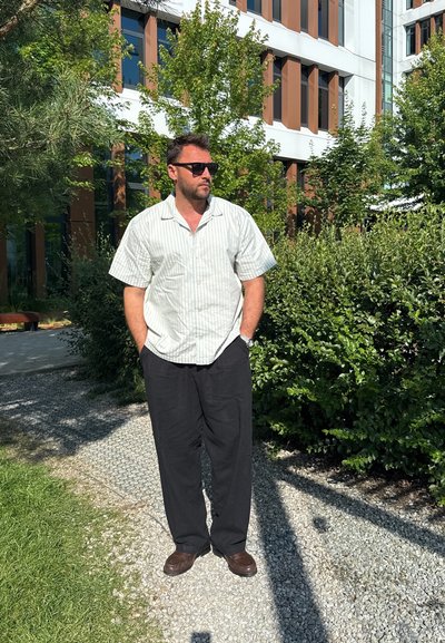 Camisa blanca de manga corta con rayas verdes, pantalones negros de corte holgado y zapatos marrones; entorno al aire libre con vegetación y fondo de edificio.