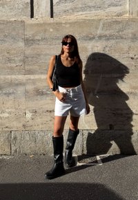 Kvinne i svart tank top, hvite shorts og svarte støvler som står mot en teksturert steinvegg, og kaster en skarp skygge.