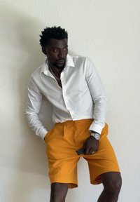 Man wearing a white button-up shirt and bright orange shorts, leaning against a white textured wall, holding sunglasses in one hand.