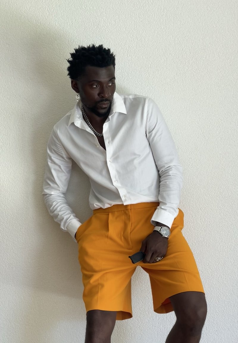 Homme portant une chemise blanche à boutons et un short orange vif, appuyé contre un mur blanc texturé, tenant des lunettes de soleil dans une main.