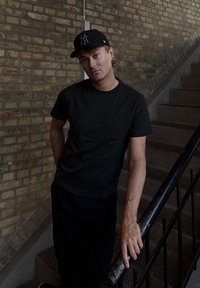 Zwart T-shirt met korte mouwen en zwarte broek gedragen door een persoon die tegen een trapleuning leunt in een omgeving met een gestructureerde bakstenen muur.