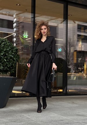 Vestido de satén negro con mangas largas, un corte holgado y cintura ceñida. Combinado con accesorios negros y medias transparentes hasta los tobillos.