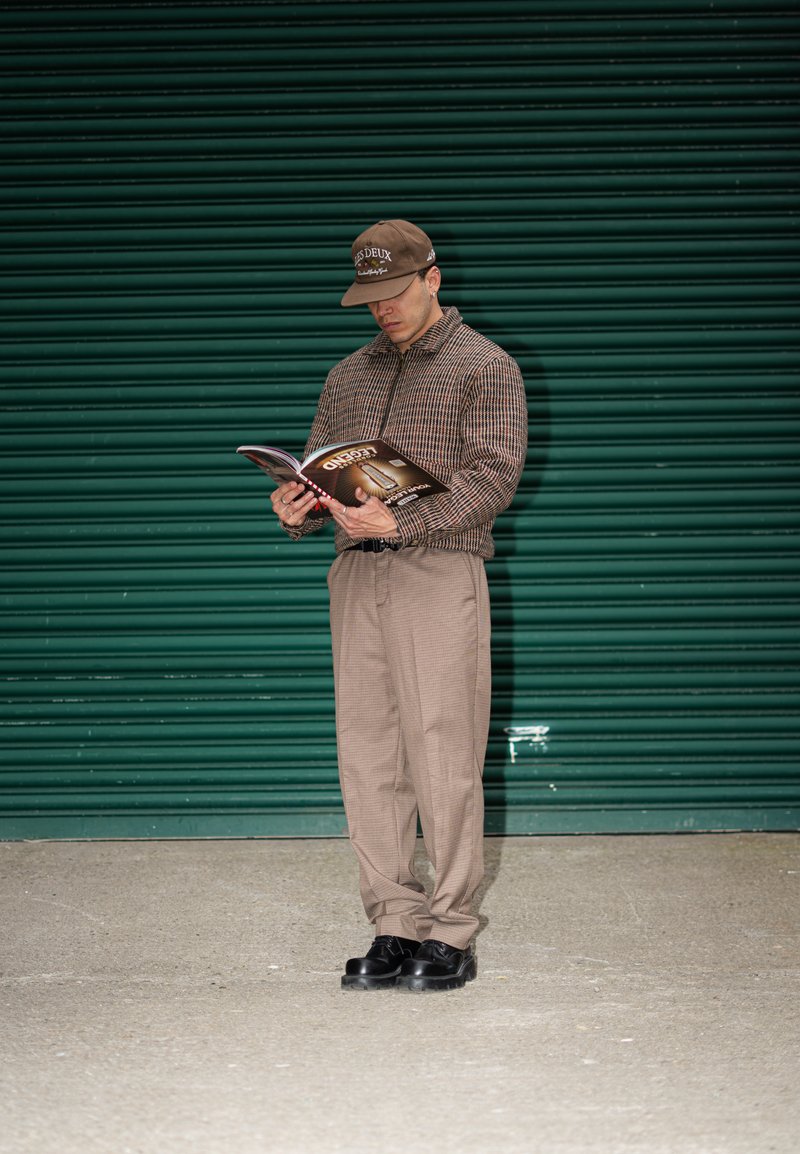 Brown checked zip-up jacket with beige plaid trousers, holding a magazine. Black shoes enhance the neutral colour palette against a green background.