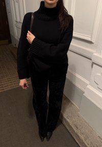 Pull à col roulé en maille noire avec poignets côtelés, associé à un pantalon large en velours noir et des talons pointus noirs, sur un fond atténué.