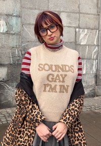 Άτομο με κοντά, ριγωτά μαλλιά που φοράει μεγάλα γυαλιά, γιλέκο πουλόβερ με τη φράση «SOUNDS GAY I'M IN», ριγωτό πουκάμισο, παλτό με λεοπάρ μοτίβο και καρό φούστα.