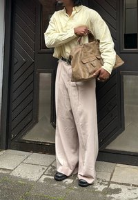 Borsa in pelle beige con due manici e una grande tasca frontale. Camicia a maniche lunghe gialla chiaro infilata in pantaloni larghi crema. Scarpe nere.