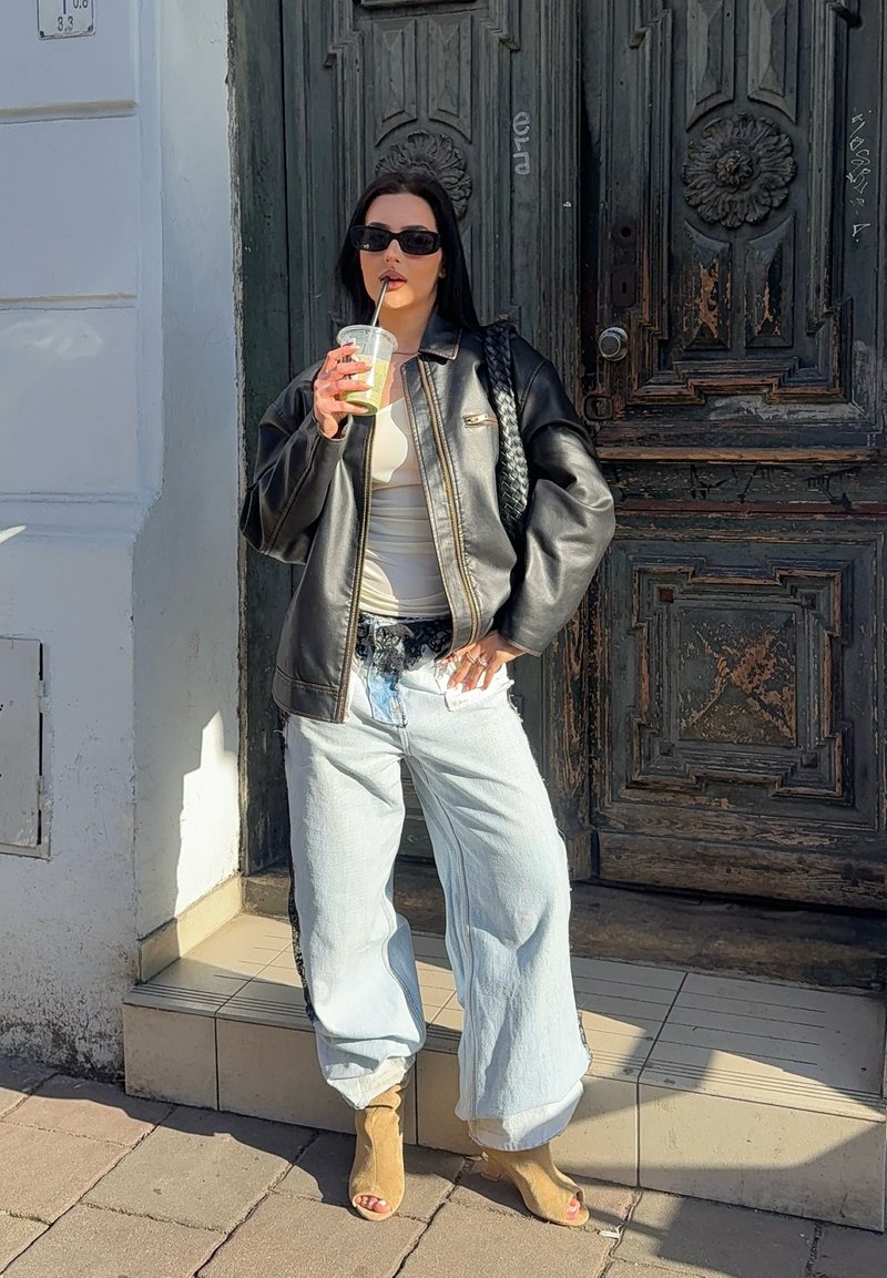 Femme portant une veste en cuir noire, un pantalon large bleu clair et des lunettes de soleil, buvant dans un gobelet en plastique avec une paille, debout près d'une porte en bois ornée.