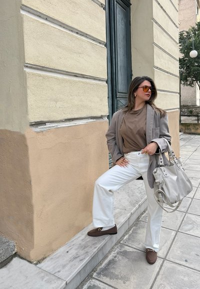 Mujer con gafas de sol, blazer gris, camisa marrón, pantalones blancos y mocasines marrones, de pie en la acera de la ciudad, sosteniendo un gran bolso gris.