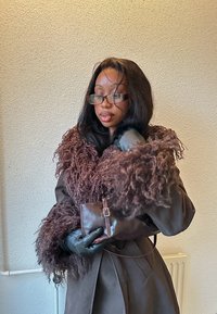 Femme portant des lunettes et un manteau marron bordé de fourrure tient une petite pochette en cuir avec des mains gantées contre un mur beige.