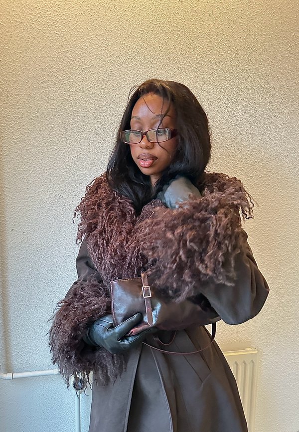 Mujer con gafas y un abrigo marrón forrado de piel sostiene un pequeño bolso de cuero con guantes contra una pared beige.