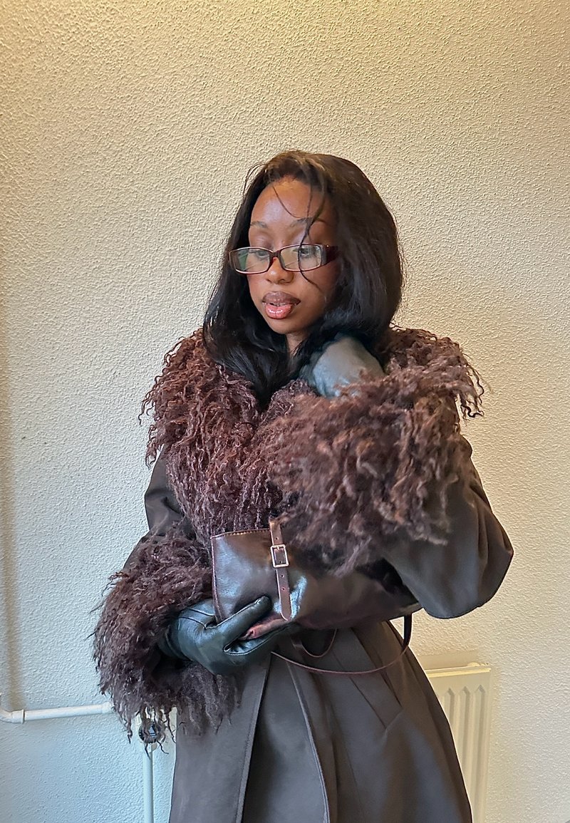 Frau mit Brille und braunem, mit Pelz gefüttertem Mantel hält mit Handschuhen eine kleine Lederclutch an eine beige Wand.