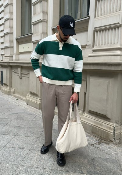 Suéter a rayas verdes y blancas con cuello, pantalones beiges, zapatos negros y un bolso de mano color crema, con un fondo de edificio de piedra.