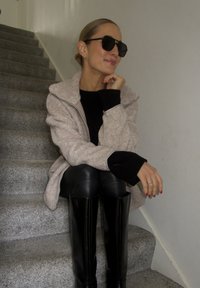 Manteau beige clair en peluche avec col, pull noir et pantalon en cuir noir ajusté. Bottes noires montantes jusqu'aux genoux et lunettes de soleil foncées. Texture lisse.