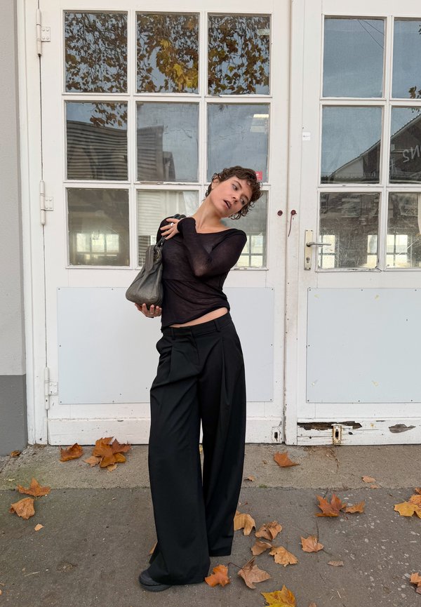 Black sheer long-sleeve top paired with loose, high-waisted black trousers. Accessories include a grey handbag. Background features a door and fallen leaves.