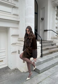 Rudos spalvos dirbtinio kailio paltas su tekstūruotomis rankovėmis, derinamas su nėriniuotomis rudomis pėdkelnėmis ir baltais aukštakulniais stiletto, fone matosi akmeninės laiptinės.