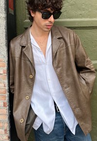 Veste en cuir marron avec coupe décontractée, boutons sur le devant, portée sur une chemise blanche ; accompagnée de lunettes de soleil foncées et d'un jean en denim.