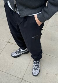 Calças pretas da Nike com bolsos laterais e um logótipo prateado, combinadas com ténis cinza e pretos com padrão, com solas grossas e atacadores.