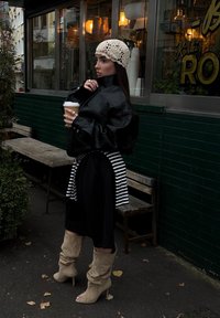 Casaco oversized de couro preto, calças largas pretas, camisa às riscas presa na cintura, botas bege até ao joelho, segurando um copo de café.