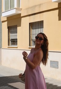 Απαλή ροζ καλοκαιρινή φόρεμα με λεπτές τιράντες, υφασμάτινη υφή, στρωτό κολιέ και γυαλιά ηλίου. Φοριέται με μια κρέμα τσάντα, μπροστά από τον κίτρινο τοίχο.