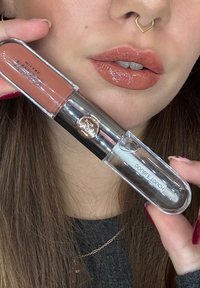 Kiiltävää alastonruskeaa huulikiiltoa levitetty huulille, joita pidellään suun lähellä; nenässä näkyy kultainen septum-renka.