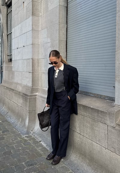 Un blazer negro ajustado sobre una camisa de rayas con cuello, combinado con unos pantalones sueltos azul marino y zapatos oscuros. Completando el look hay un tote negro tejido.