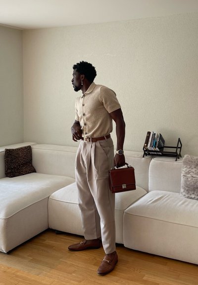 Hombre con polo de punto beige y pantalones plisados sosteniendo un maletín marrón, de pie junto a un sofá seccional blanco con cojines.