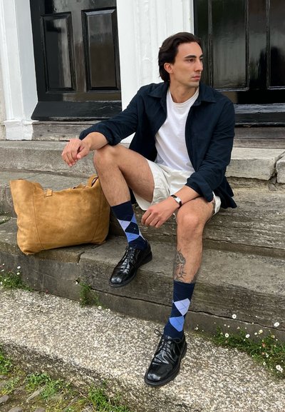 Zapatos de cuero negros, pantalones cortos color crema, camisa azul marino, camiseta blanca, calcetines de rombos en tonos azules y un bolso de ante color canela.