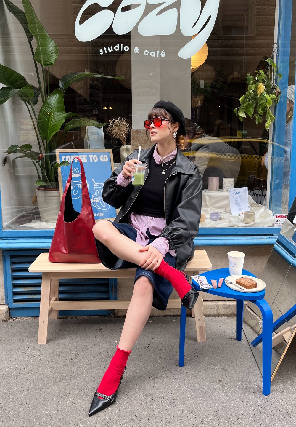 Κόκκινη δερμάτινη τσάντα, μαύρο δερμάτινο μπουφάν, ροζ πουκάμισο, σκούρα τζιν φούστα, κόκκινες κάλτσες, μαύρες μυτερές γόβες. Μπλε και μπεζ καφετέρια με φυτά.