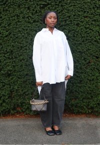 Biała, oversize koszula na guziki, ciemnoszare workowate dżinsy, czarne baleriny oraz mała czarna torebka z złotym łańcuszkiem. Zielone tło.